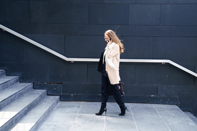 Free photo business woman in a coat with a bag in her hands goes up the steps to the building. the concept of career and business