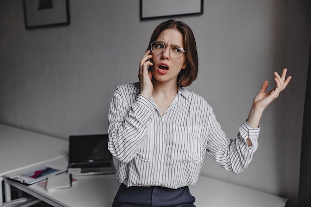 Free Photo business woman in stylish blouse emotionally talking on phone. shot of girl with glasses on background of white table with stationery.