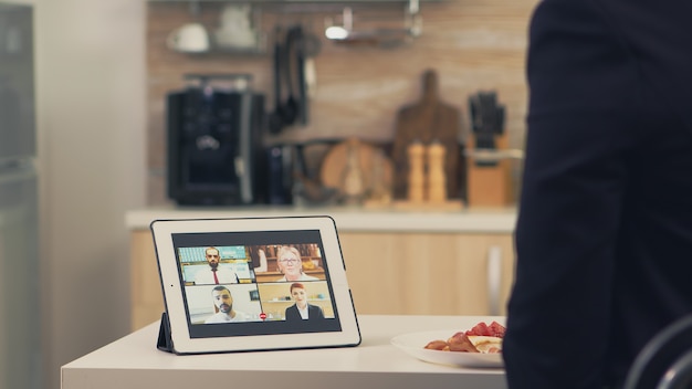 Business woman using smart device while eating breakfast in kitchen. Young freelancer at home talking on a video call with her colleagues from the office, using modern internet technology