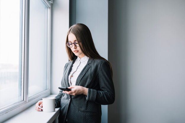 Business woman with coffee using smartphone at window 