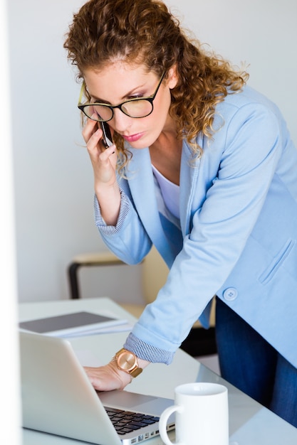 Free Photo business young woman talking on the mobile phone while using her laptop in the office.