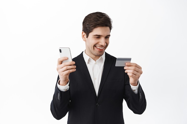 Businessman in black suit look at credit card while paying online with mobile phone shopping online on smartphone app order something contactless white background