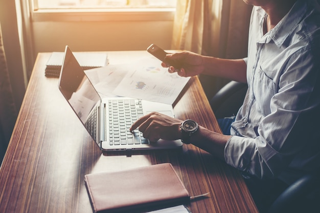 Businessman hands using cell phone with laptop at office desk.