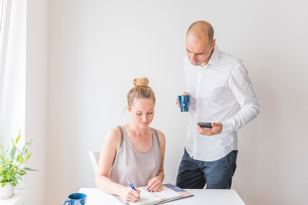 Businessman holding calculator and coffee cup looking at woman writing in diary