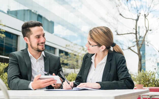 Businessman holding mobile in hand looking at her colleague writing on the document