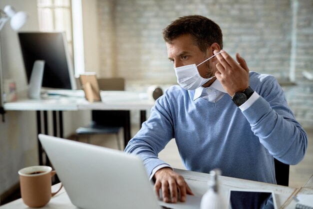 Businessman putting on protective face mask while working on laptop in the office