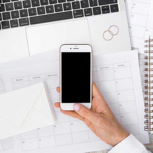 Free Photo businessman's hand holding smartphone over the calendar with envelope; laptop and wedding rings