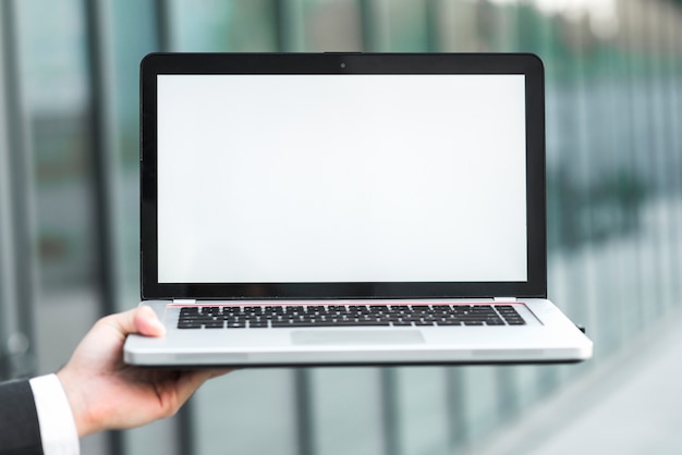 Free photo a businessman's hand showing laptop with blank white screen against blurred backdrop