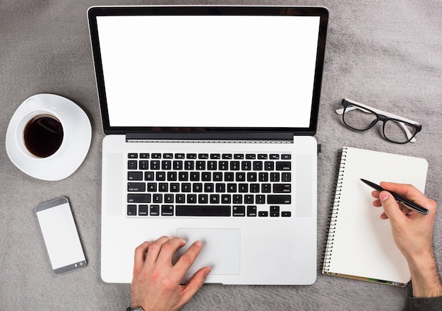 Free photo a businessman's hand using digital tablet writing on spiral notepad over the gray desk