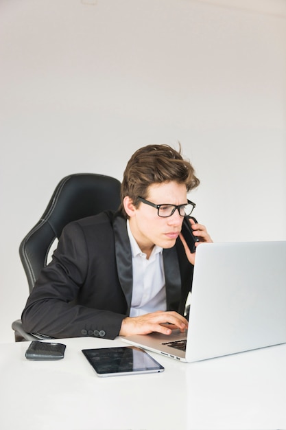 Businessman talking on phone while using laptop at workplace