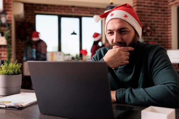 Free photo businessman working on laptop in decorated office with christmas tree and festive ornaments. male worker using pc in workplace with xmas decorations during winter holiday season.