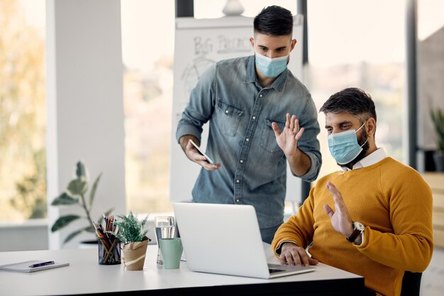 Businessmen with face masks waving during video call over laptop in the office