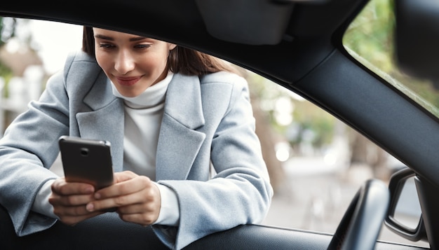 Businesswoman leaning on car window and texting message on phone, smiling happy.