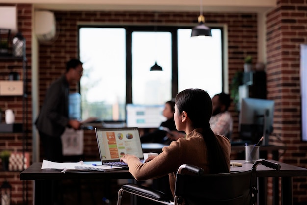 Businesswoman living with disability analyzing report on laptop, working with statistics in disability friendly office. Asian employee suffering from chronic impairment, using wheelchair.