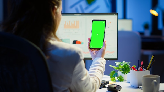 Free photo businesswoman looking at green screen monitor of smartphone sitting at desk in business office late at night. freelancer watching desktop monitor display with green mockup, chroma key working overtime