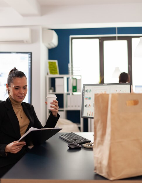 Businesswoman sitting at desk in corporate office reading financial statistics on clipboard, before enjoying tasty delicous food from takeaway in paper bag