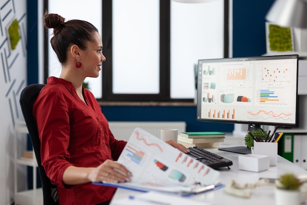 Businesswoman with clipboard sitting at desk in startup business office. Smiling employee in red shirt comparing charts. Successful entrepreneur looking at desktop computer to analyze business data.