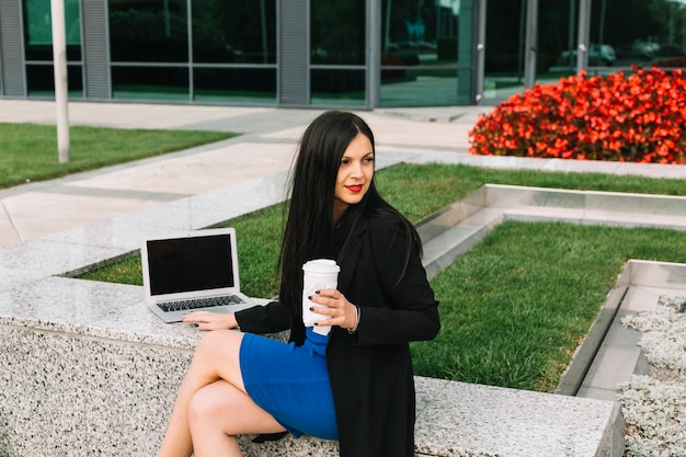 Businesswoman with laptop and disposal cup sitting outside office building