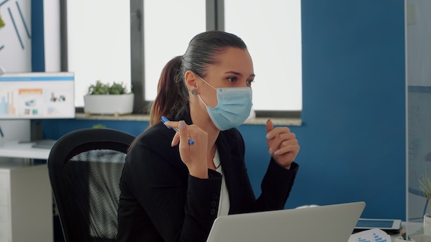 Businesswoman with medical face mask talking with her team about communication strategy sitting at desk. Business team keeping ing ing social distancing while working in new normal company office