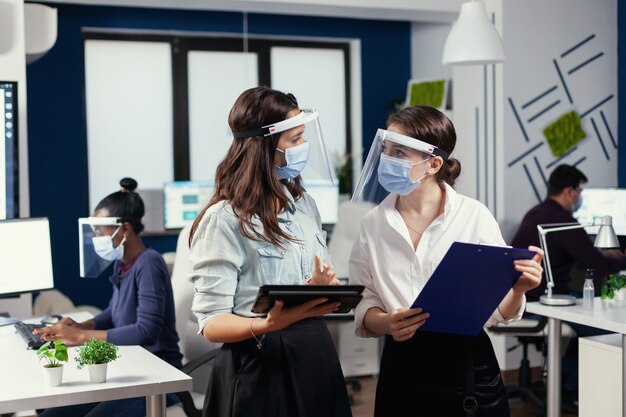 Businesswomen with protection masks against coronavirus standing in workplace talking about company financial datas holding digital tablet. Multiethnic business team working respecting social distance