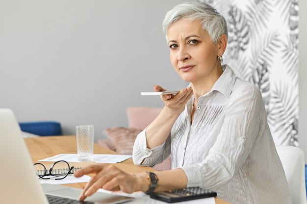 Busy attractive gray haired Caucasian woman accountant on retirement working as freelancer managing finances, sitting at desk with portable computer, holding mobile phone, recording voice message