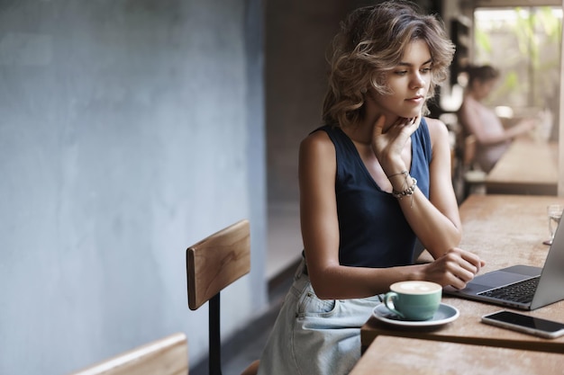 Free photo busy attractive young modern businesswoman prepare project reading article sit near window cafe table drink coffee thinking look laptop display female student tutor make arrangements future lesson