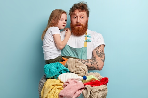 Busy foxy single dad tries to tease crying child, looks with puzzled expression, wears apron, washes laundry, has much domestic work, isolated over blue wall. Fatherhood and business concept