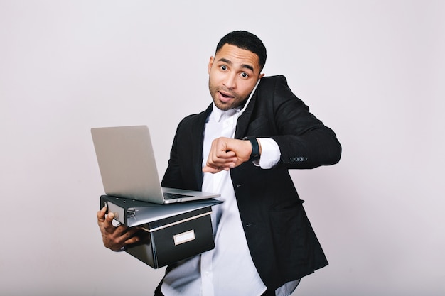 Free photo busy hard-working guy in white shirt and black jacket talking on phone, holding folders, laptop, looks astonished at watch. office worker, modern technology, career.