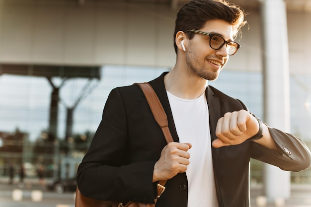 Busy man looks at watch and smiles Portrait of brunette handsome guy in black jacket and white tshirt holding brown backpack Businessman in eyeglasses poses outside