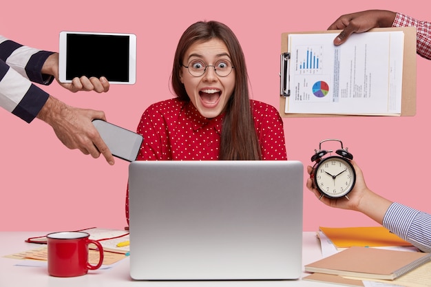 Busy woman wears spectacles, being overworked, has deadline for preparing task, surrounded with laptop, hands hold electronic gadgets