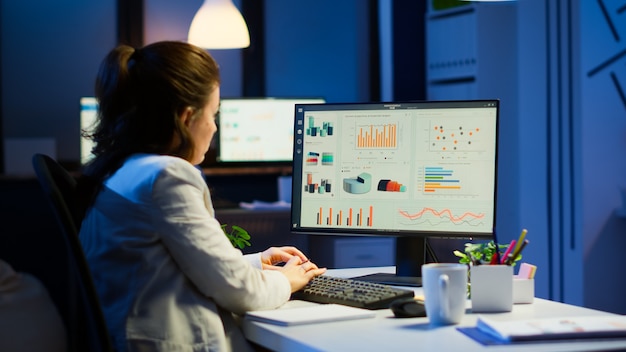 Busy woman working at night in front of computer taking notes writing on notebook annual reports, checking financial project. Focused employee using technology network wireless doing overtime for job