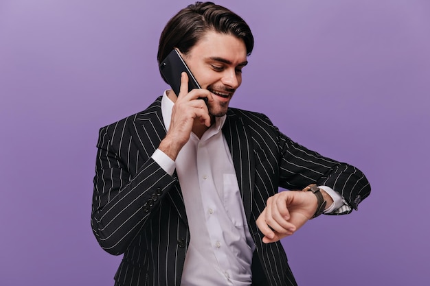 Busy young gentleman with brunette hair white shirt and black striped suit talking on phone looking at watch and smiling against violet background Business concept