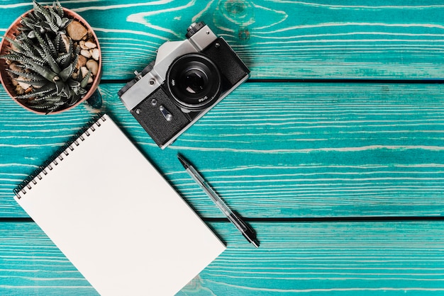 Free Photo cactus pot plant; camera; spiral note pad and pen on turquoise wooden plank