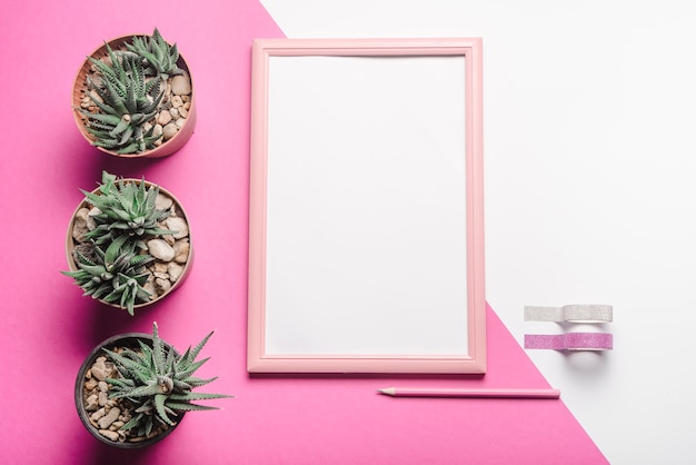 Free photo cactus potted plant; white blank frame; pencil and ribbons on white and pink background