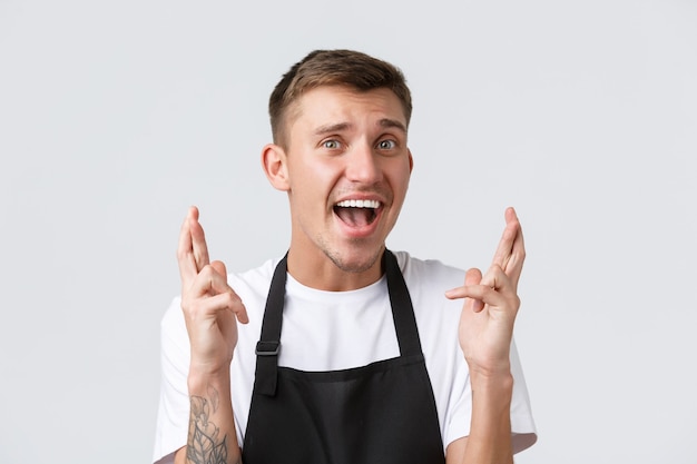 Cafe and restaurants, coffee shop owners and retail concept. Hopeful and excited handsome waiter in black apron, cross fingers good luck and looking amazed camera, praying over white background