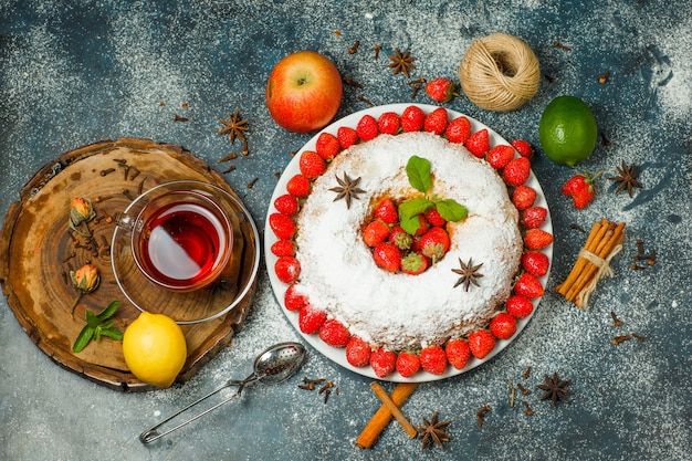 Cake with fruits, strainer, tea, thread, spices, sugar, herbs in a plate on wooden board and stucco background, top view.