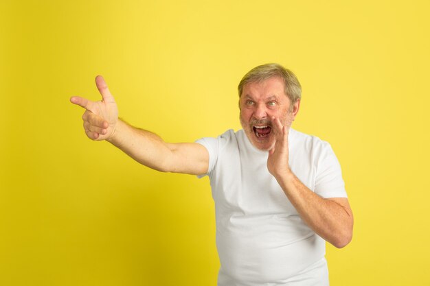 Calling, pointing on. Caucasian man portrait isolated on yellow studio background. Beautiful male model in white shirt posing. Concept of human emotions, facial expression, sales, ad. Copyspace.