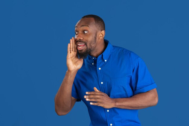 Calling, shouting. Monochrome portrait of young african-american man isolated on blue studio wall.