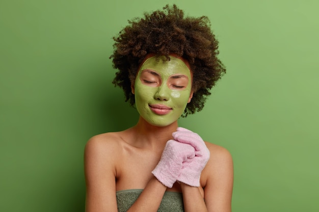 Free Photo calm afro american with curly bushy hair holds bath gloves wrapped in soft towel applies green moisturizing mask