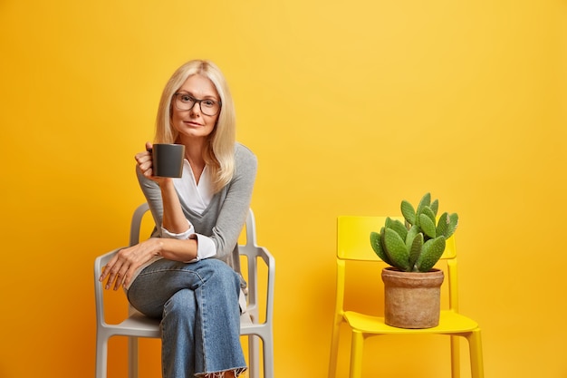 Free photo calm blonde woman sits on comfortable chair with beverage looks confidently and poses near potted cactus enjoys quiet atmosphere. lifestyle concept