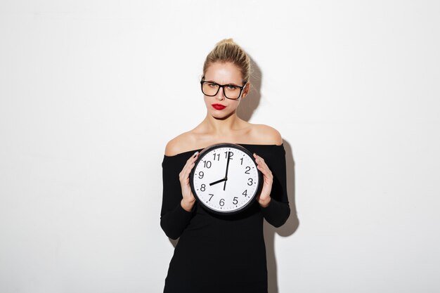 Calm business woman in dress and eyeglasses holding clock