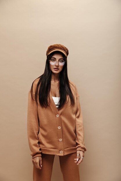 Calm serious woman in light brown stylish cashmere suit and cap looks into camera Brunette longhaired girl in eyeglasses poses on isolated beige background