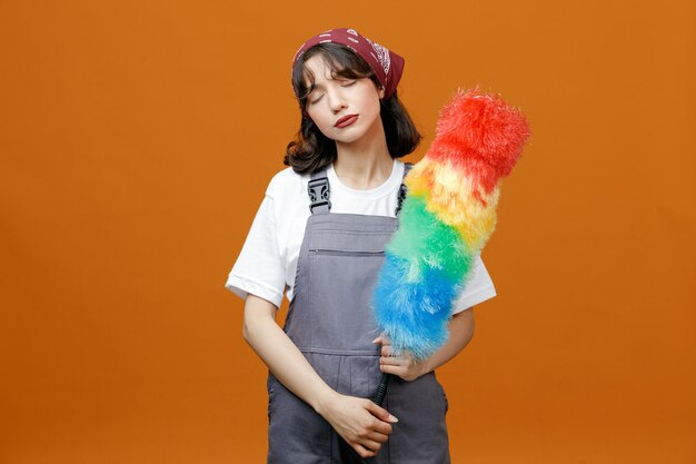 Calm young female cleaner wearing uniform and bandana holding feather duster with both hands with closed eyes isolated on orange background