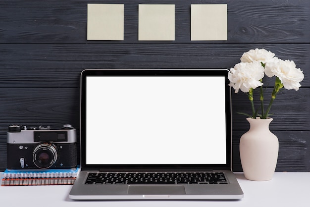 Camera; spiral notebook; flower vase and an open white blank laptop on desk against wooden background