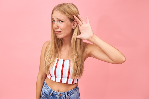 Free Photo can you speak louder? portrait of fashionable cute young female in crop summer top and jeans frowning, holding hand at her ear, having hearing problem because of noise