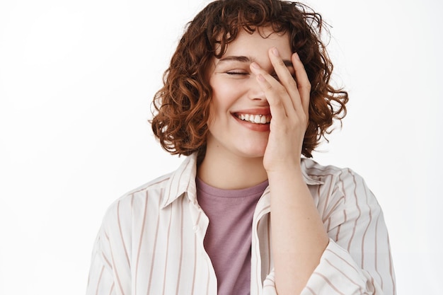 Candid happy woman with beautiful smile, covering face with hand shy, blushing while laughing and having fun, standing relaxed against white background