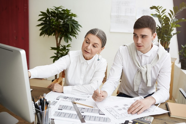 Candid shot of attractive mature female developing construction project in light office, working together with her young male colleague, using CAD system on generic computer, pointing at screen