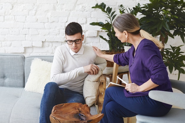 Candid shot of casually dressed professional woman psychotherapist in her fifties touching her young male patient by shoulder while having counseling session, expressing sympathy and support