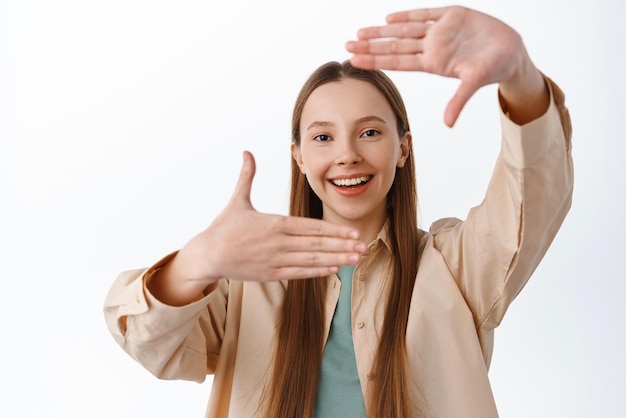 Free photo capture moment smiling young girl making hands frames camera gesture and looking through searching for perfect angle imaging something standing over white background