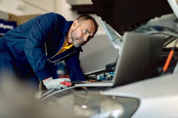 Car mechanic using laptop while examining engine in auto repair shop